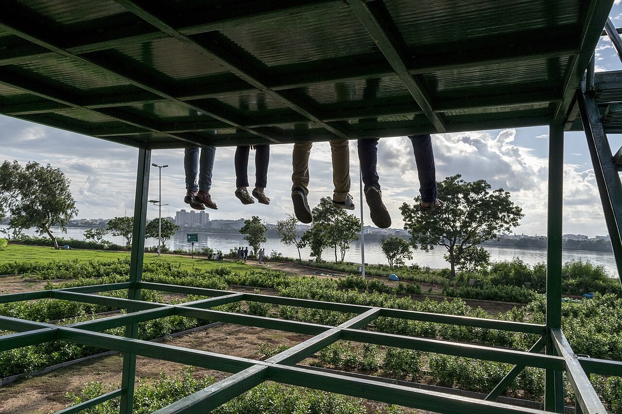 Piernas de cuatro amigos sentados en una estructura elevada, junto a un parque y un lago; sus rostros quedan fuera del encuadre.
